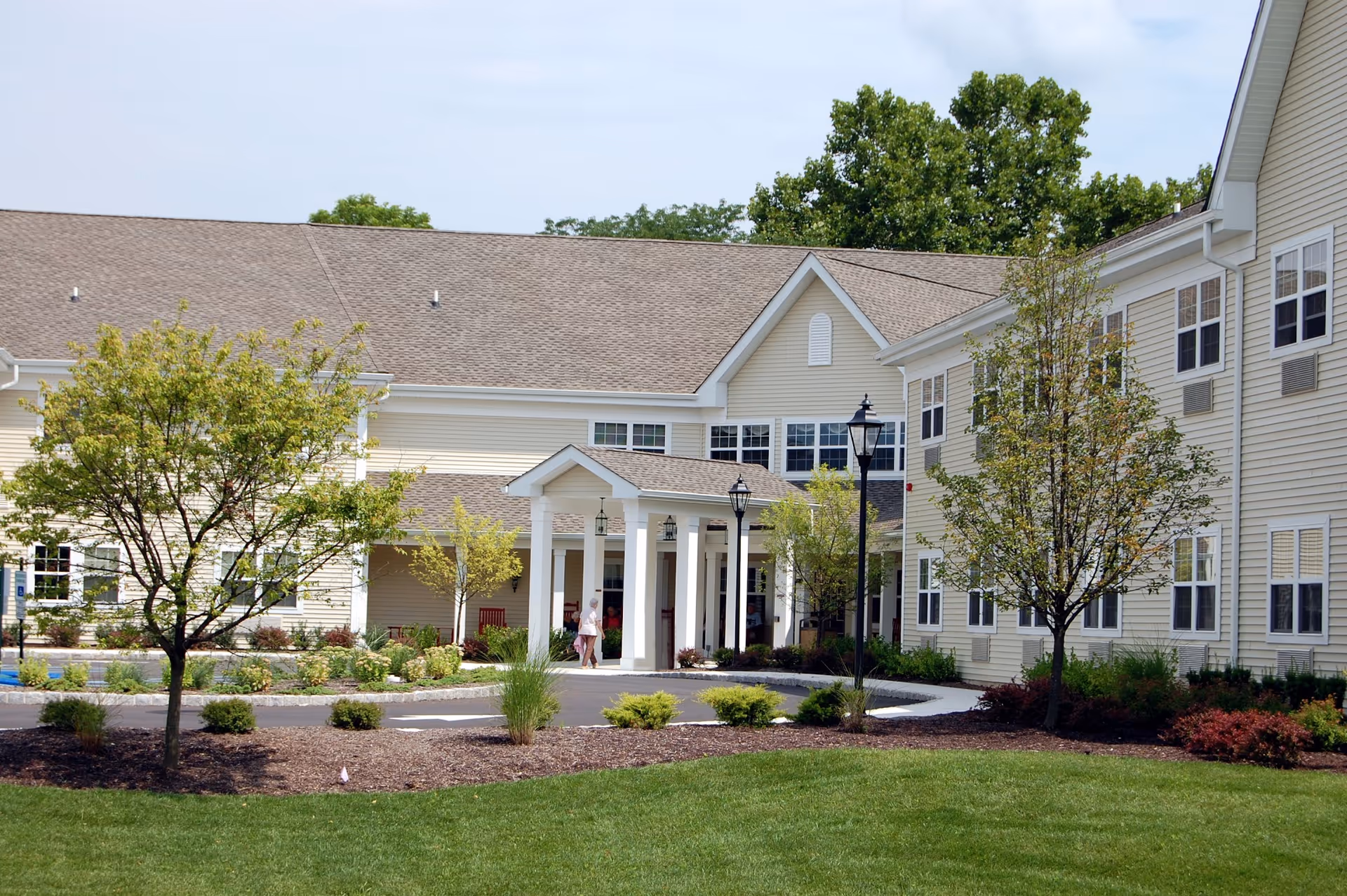 Front entrance of a beige senior living facility with a covered portico, landscaped lawn, and trees.