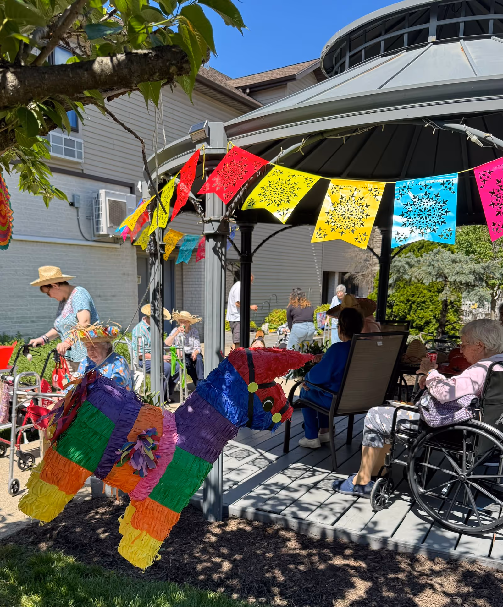 Outdoor gathering at a senior living facility with elderly people sitting under a gazebo decorated with colorful paper flags. A colorful piñata shaped like a donkey hangs nearby. Some seniors are seated in wheelchairs and others are using walkers. The weather is sunny and the setting is festive.