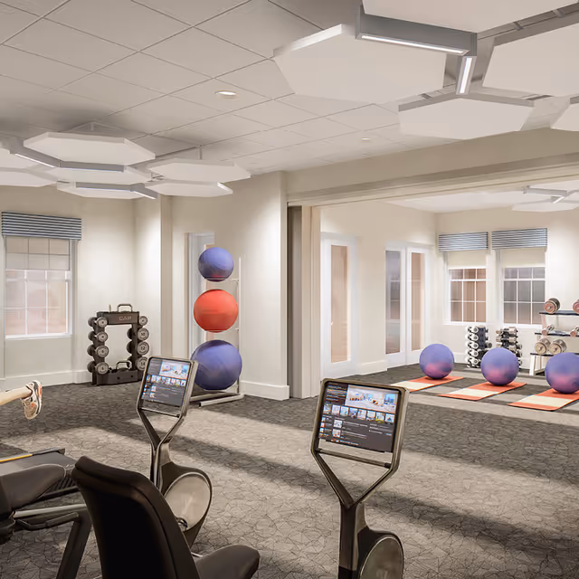 A bright fitness room with stationary bikes featuring screens, exercise balls, dumbbells, and yoga mats.