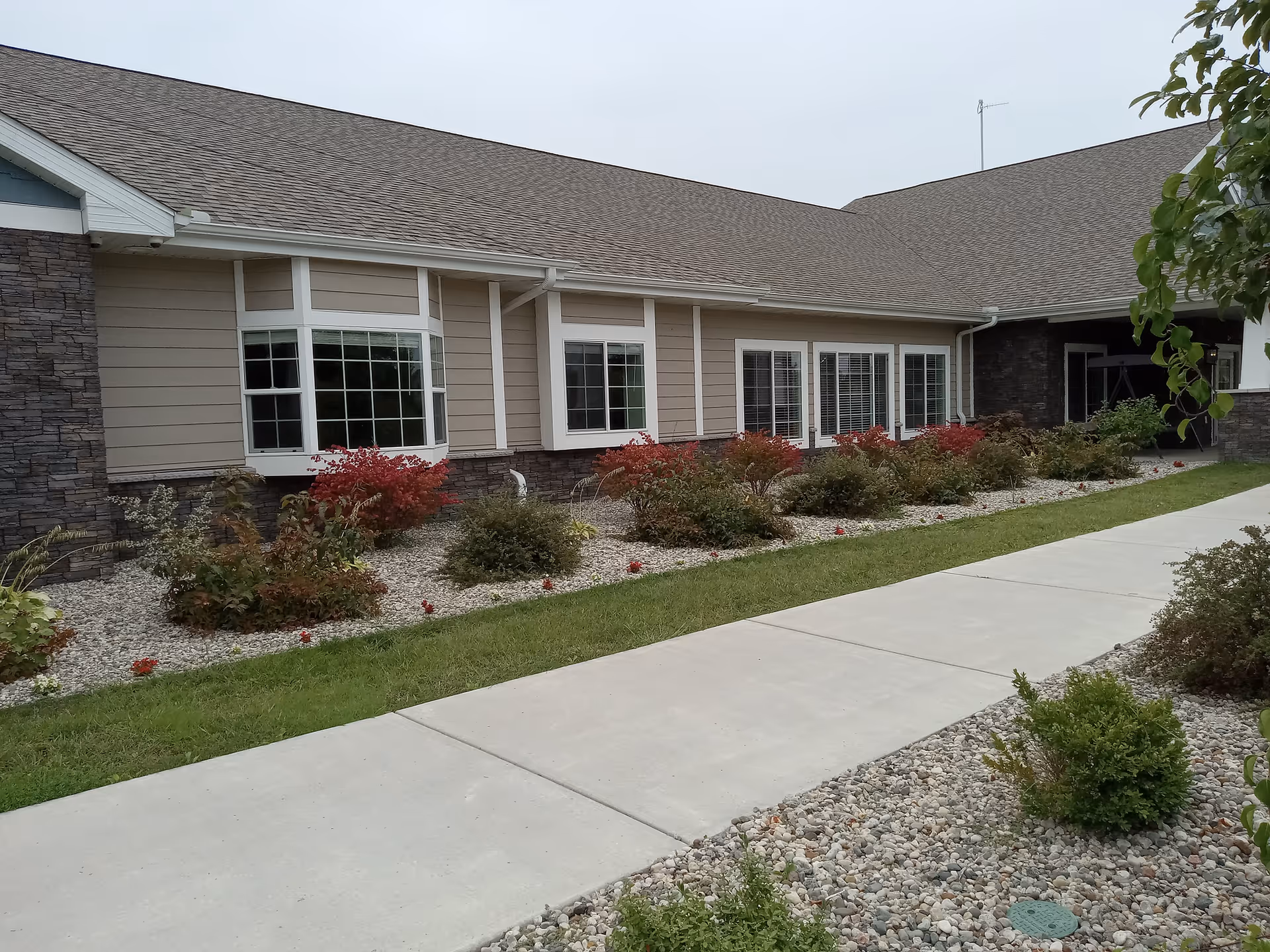 Exterior view of a single-story assisted living facility building with beige siding and stone accents. There are several windows with white frames and a landscaped area with bushes and small plants along a concrete walkway.