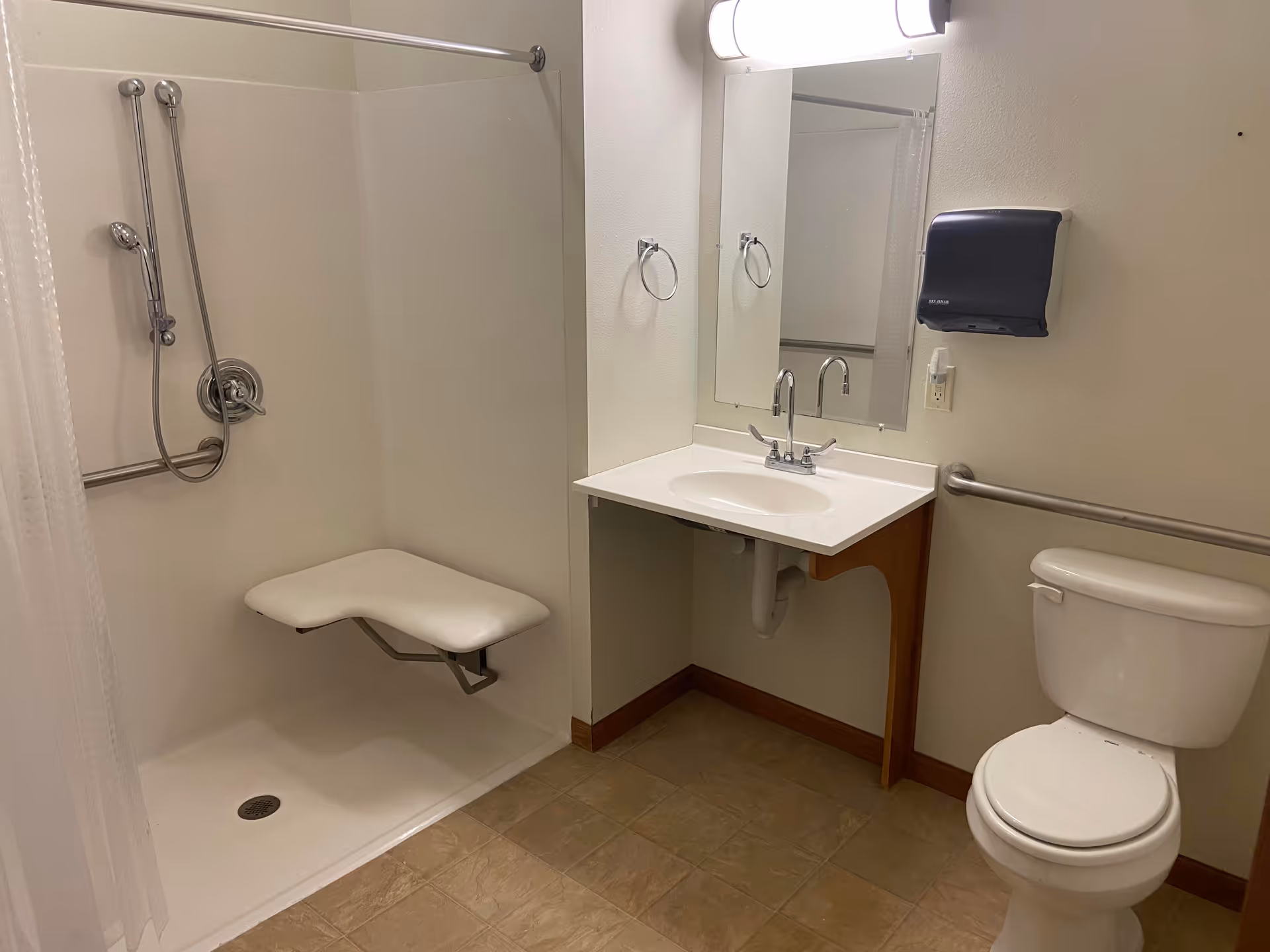 A clean, accessible bathroom featuring a walk-in shower with a fold-down seat and handheld showerhead, a white sink with a mirror above it, a toilet, and grab bars along the walls. The floor is tiled and the walls are painted a light color.