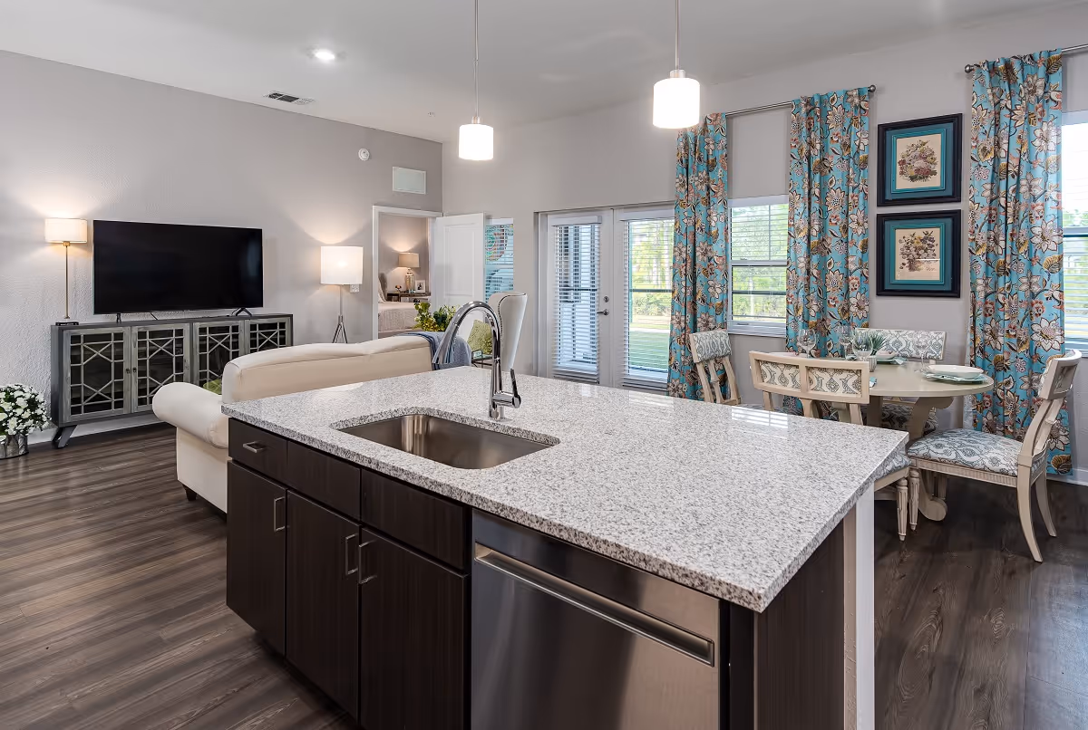 Modern open-concept living space featuring a kitchen island with a sink and granite countertop in the foreground, a beige sofa facing a flat-screen TV on a decorative cabinet, and a dining area with a round table and four chairs near windows with floral blue curtains.