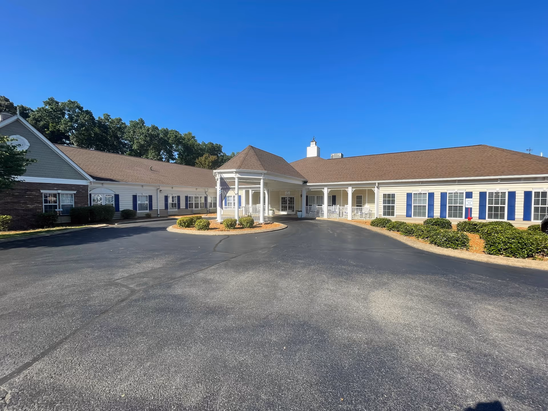 Exterior view of a single-story senior living facility building with a covered entrance and a circular driveway. The building has beige siding with blue shutters and a brown roof. There are bushes and small landscaped areas around the driveway, and trees are visible in the background under a clear blue sky.