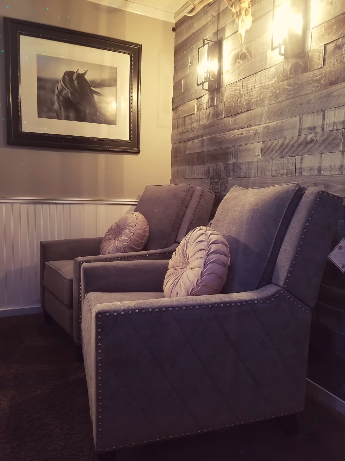 Two gray upholstered armchairs with round pink cushions are placed side by side in a cozy corner. The wall behind the chairs is covered with wooden panels and has two lit wall sconces. A framed black and white picture of a horse hangs on the adjacent wall.