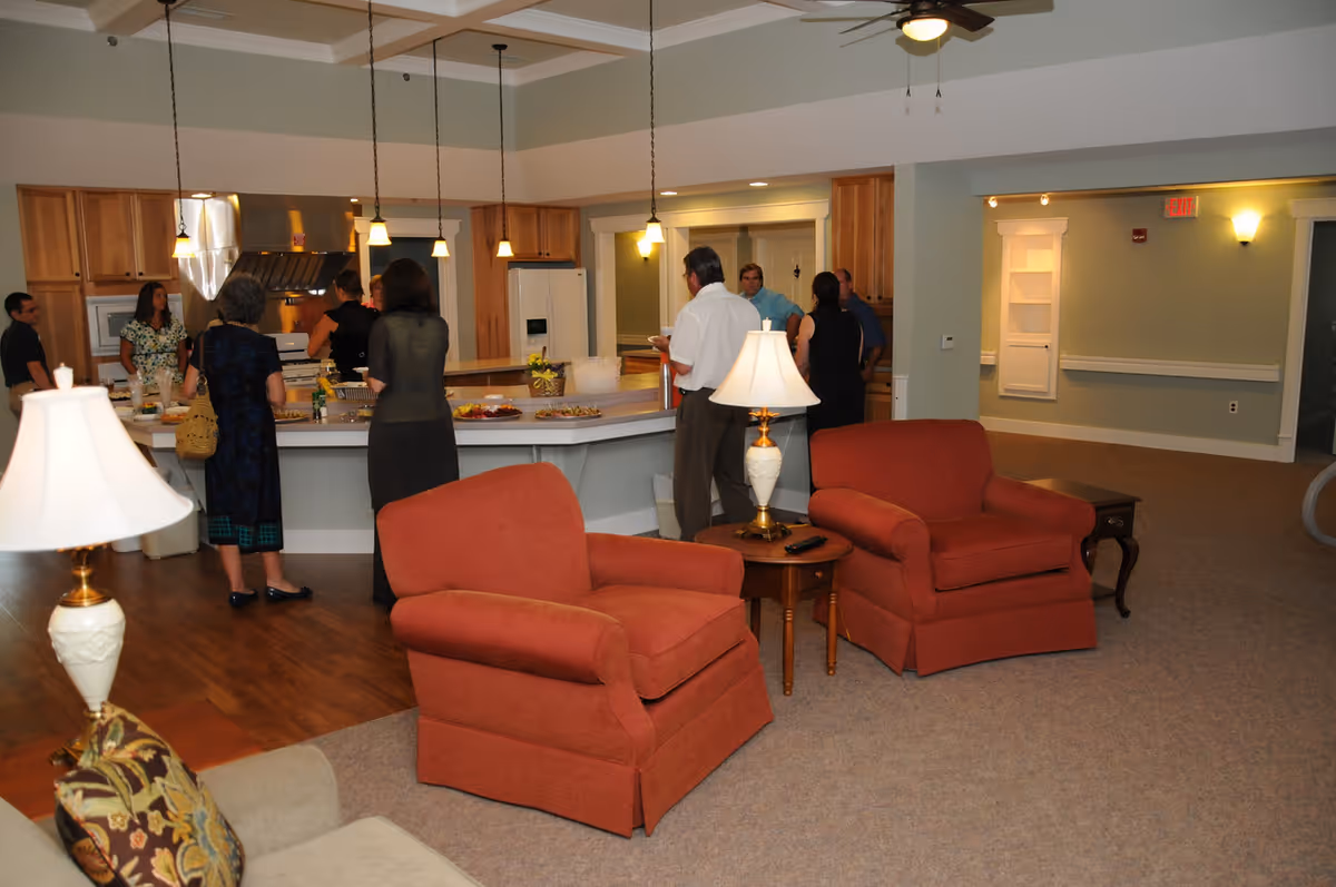 A group of people gathered in a senior living facility's open kitchen and living area. The kitchen features wooden cabinets, a refrigerator, and a stove with a hood. Several pendant lights hang over the kitchen island where food is placed. In the foreground, there are two red armchairs with a small wooden table and a lamp between them. The room has a carpeted floor and ceiling fans with lights.