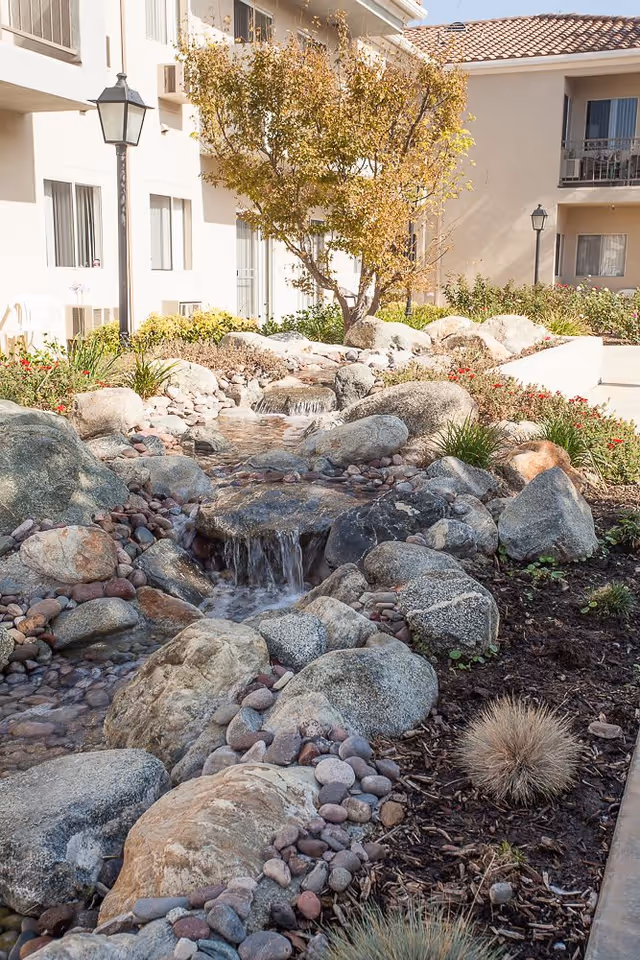A rock-lined courtyard water feature with a small cascading stream, plants, a tree, and apartment balconies in the background.