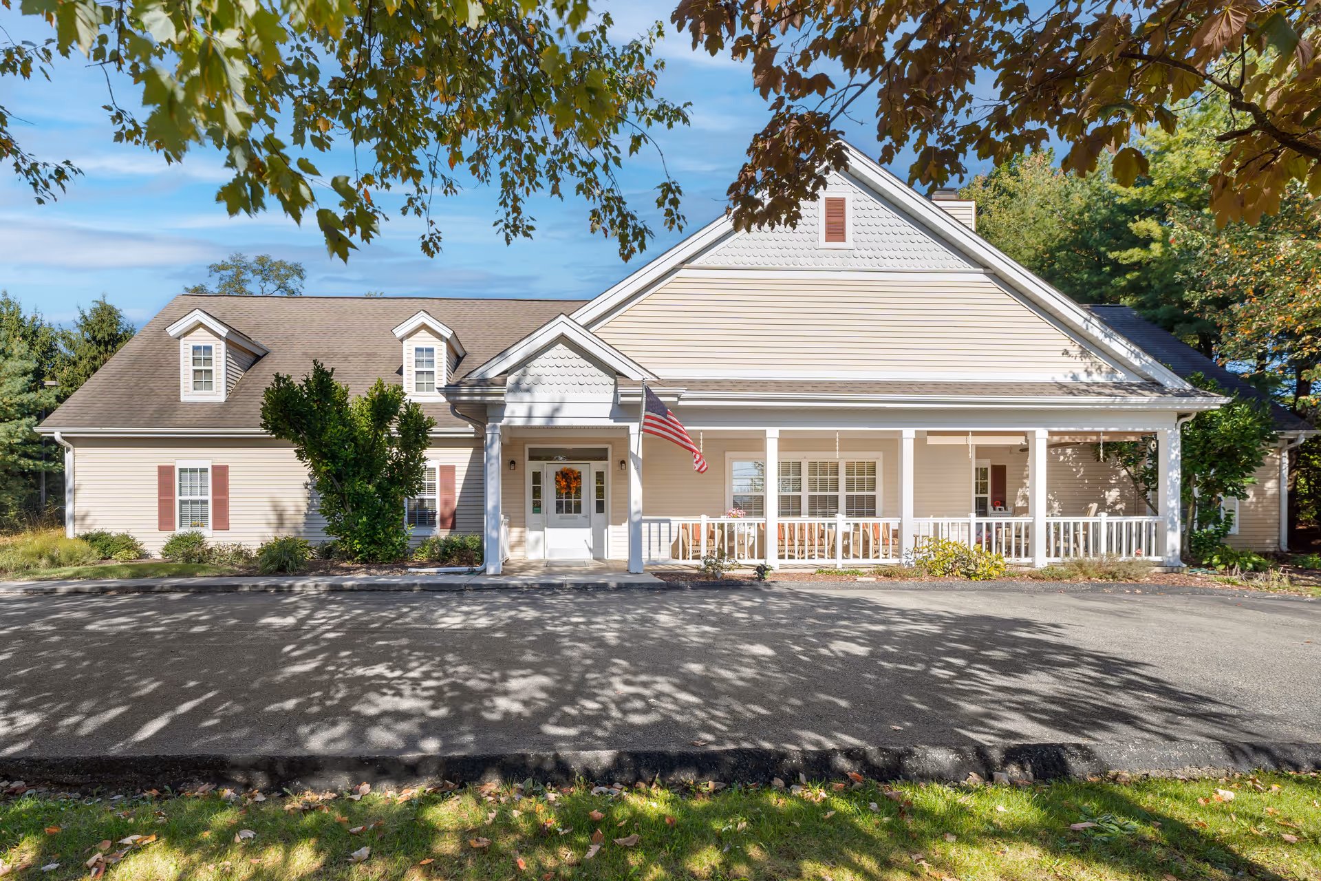 Front exterior view of a single-story beige building with a gabled roof, white columns, and a porch with seating. An American flag is mounted near the entrance door, and trees with green and brown leaves frame the top of the image.