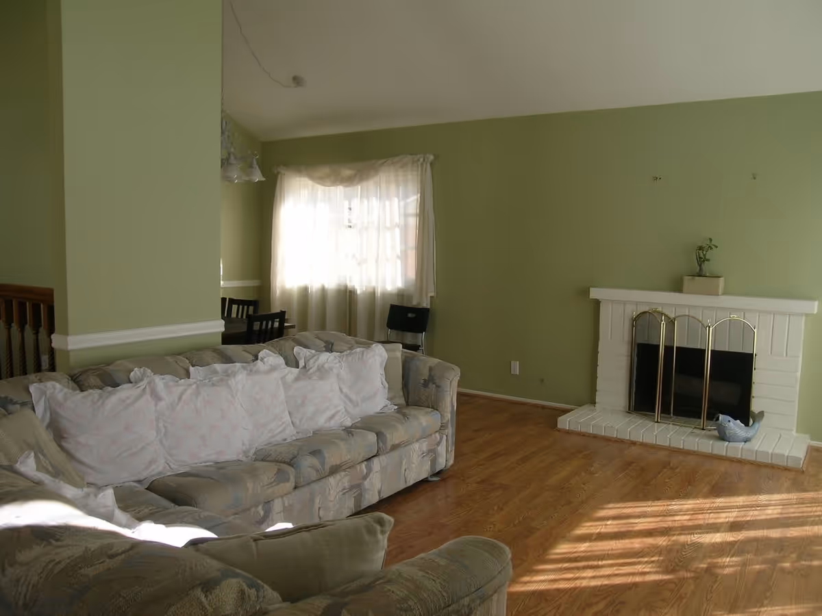 A living room with light green walls, a white brick fireplace with a brass screen, and a small plant on the mantel. There is a patterned sofa with several white pillows in the foreground, and a dining area with a table and chairs is partially visible near a window with sheer white curtains.