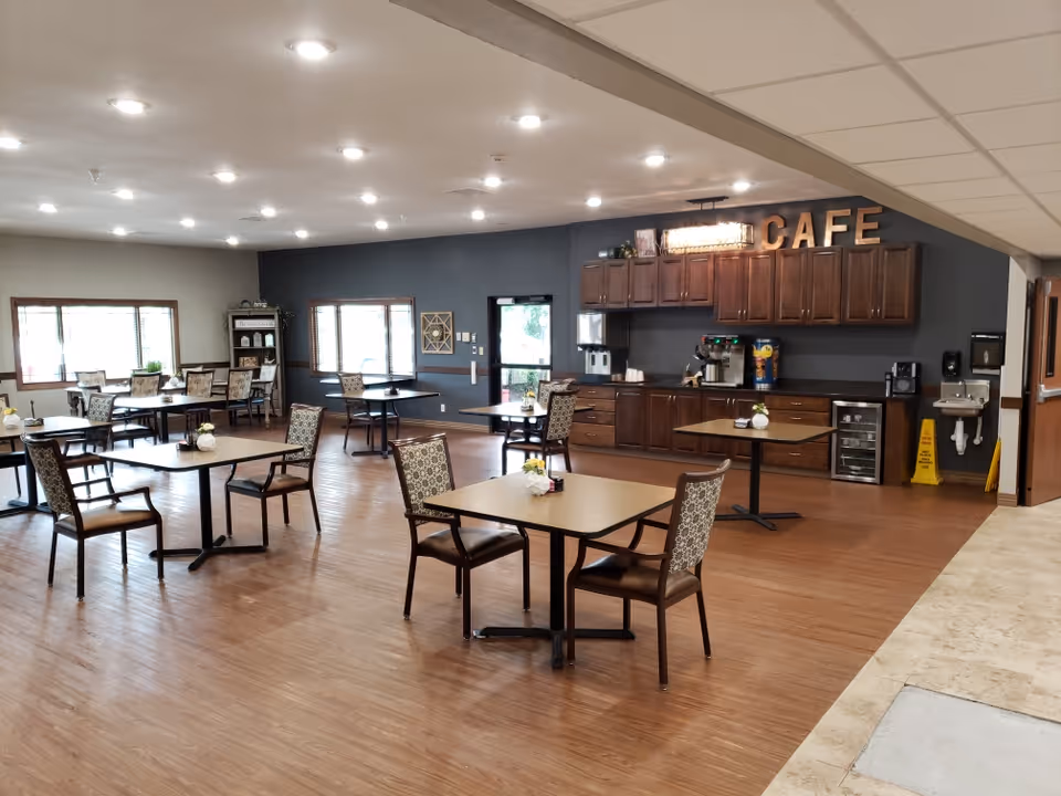 A spacious dining area with several tables and chairs arranged neatly on a wooden floor. The back wall features dark cabinetry with a coffee station and a sign that reads 'CAFE'. Large windows allow natural light to brighten the room, and there are decorative items on the tables and shelves.