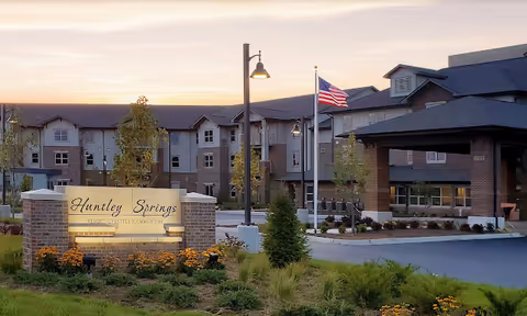 Exterior view of Huntley Springs Retirement Resort at sunset, showing the main building with multiple windows, an American flag on a flagpole, street lamps, and a landscaped area with flowers and greenery in front of a brick sign displaying the facility's name.