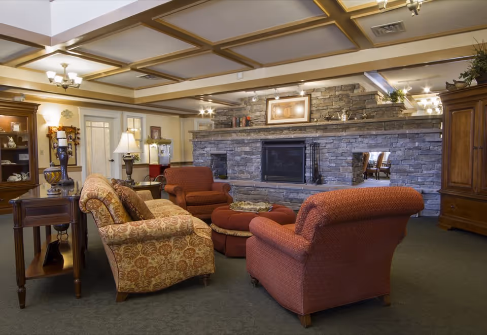 A cozy living room area in a senior living facility featuring a stone fireplace with a framed picture above it. The room has a patterned sofa, two upholstered armchairs, and a round ottoman in the center. There are wooden side tables with lamps and decorative items, and the ceiling has wooden beams with recessed lighting. The overall ambiance is warm and inviting.