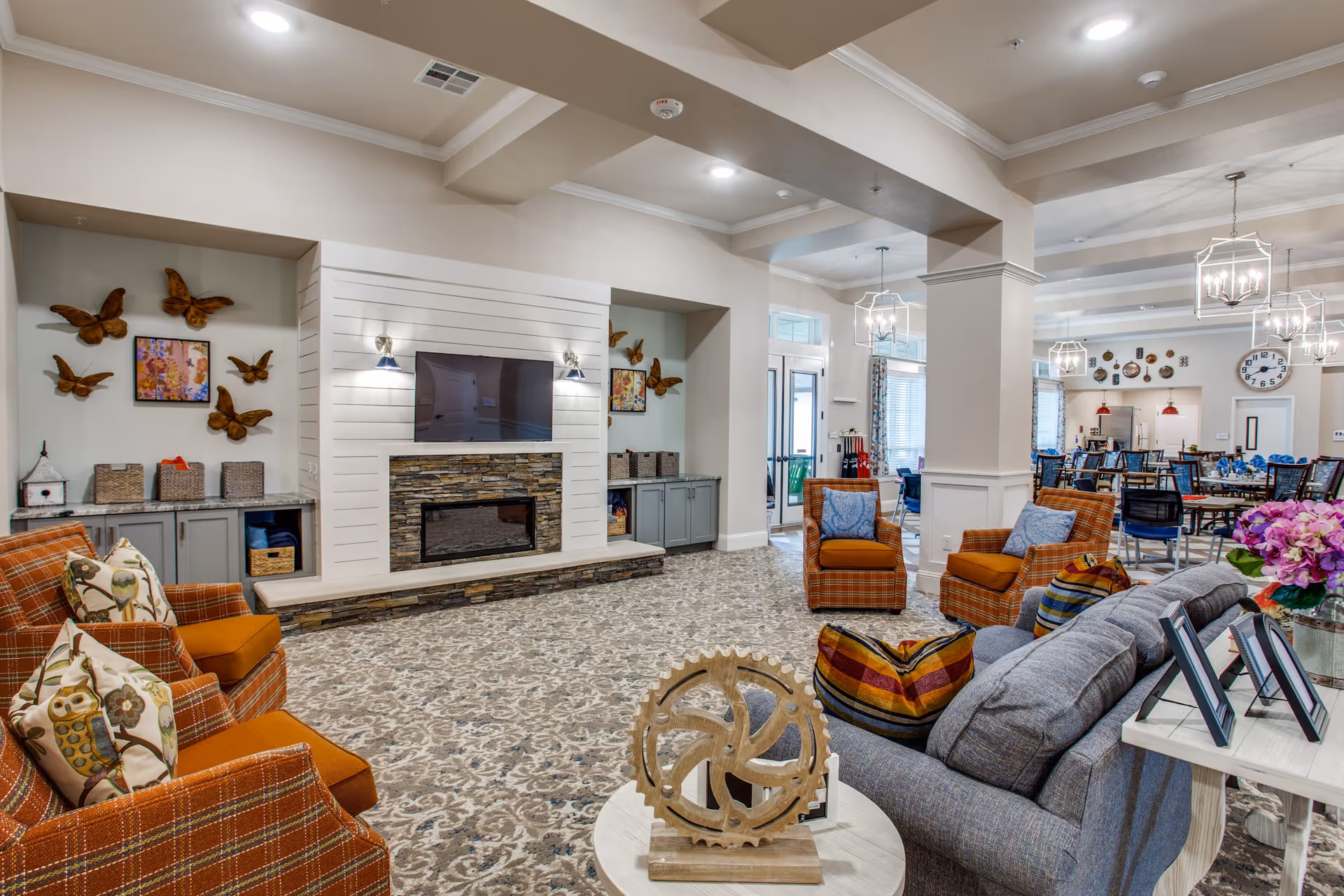 A spacious and well-lit common area in The Harvest Of Aledo featuring a cozy seating arrangement with orange plaid armchairs and a gray sofa adorned with colorful pillows. The room includes a modern fireplace with a mounted TV above it, decorative wall art with butterflies, and a patterned carpet. In the background, there is a dining area with tables and chairs, pendant lighting, and large windows allowing natural light.