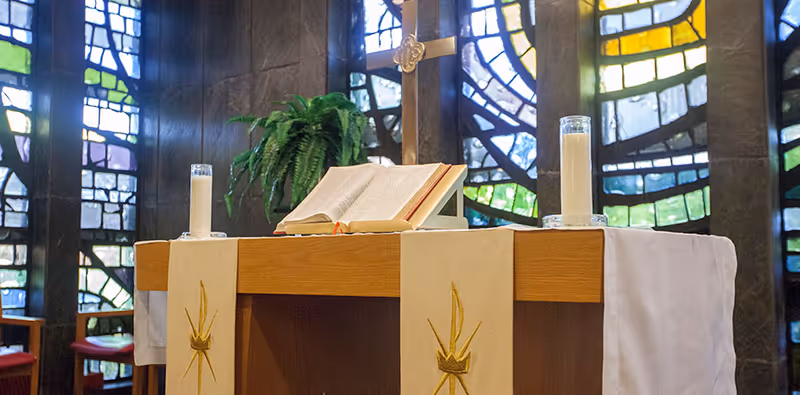 A wooden altar inside a chapel with an open Bible, two lit candles in glass holders, a gold cross, and a green plant in the background. The backdrop features colorful stained glass windows.