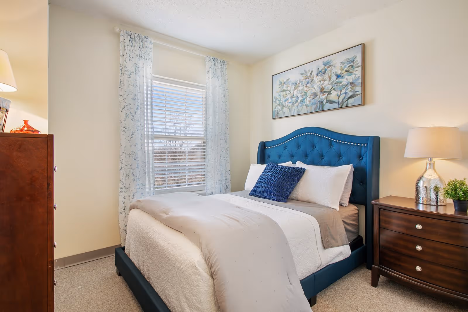 A cozy bedroom with a blue upholstered bed featuring white and blue pillows and a beige comforter. There is a window with sheer floral curtains letting in natural light. On the right side, a wooden nightstand with a silver lamp and a small green plant is visible. On the left side, a wooden dresser with a red decorative item on top is partially visible. A floral painting hangs above the bed on the cream-colored wall.