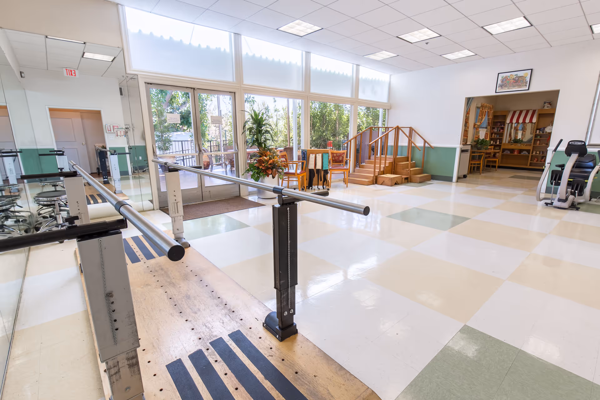 A spacious rehabilitation room with parallel bars for walking exercises, a set of wooden stairs with handrails for physical therapy, exercise equipment, large windows letting in natural light, and a small seating area with chairs and plants.