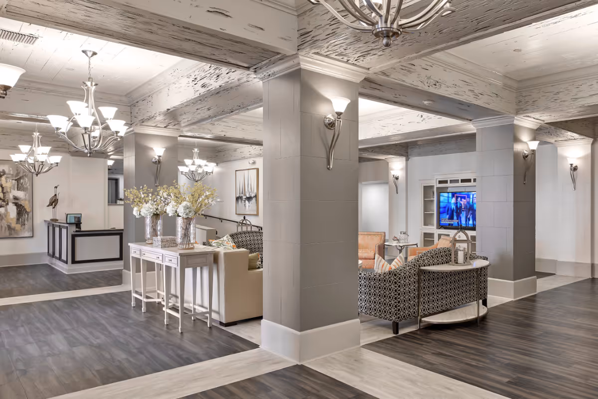 A spacious and well-lit assisted living facility common area with modern chandeliers, decorative wall sconces, and a mix of light and dark wood flooring. The room features comfortable seating arrangements including patterned sofas and armchairs around a TV, a white console table with three vases of flowers, and artwork on the walls. The ceiling has a distressed wood finish, and the overall decor is elegant and inviting.