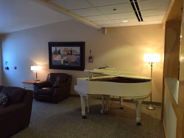 A cozy interior room featuring a white grand piano with its lid open, two brown leather armchairs, a side table with a lamp, a floor lamp, and a framed artwork on the wall depicting a scenic landscape with lighthouses and hills.