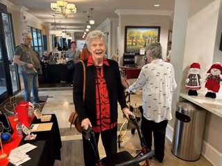 Elderly residents and staff mingling in a bright senior living facility common area with tables, holiday decorations, and a smiling woman using a walker.