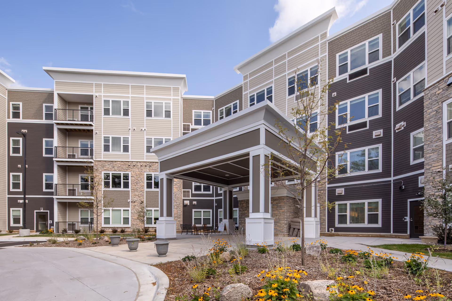Exterior view of a modern multi-story senior living facility with a covered entrance, landscaped garden with flowers and small trees, and clear blue sky.