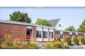 Single-story brick building with a row of windows, a small entrance with handrails, and landscaped shrubs in front.