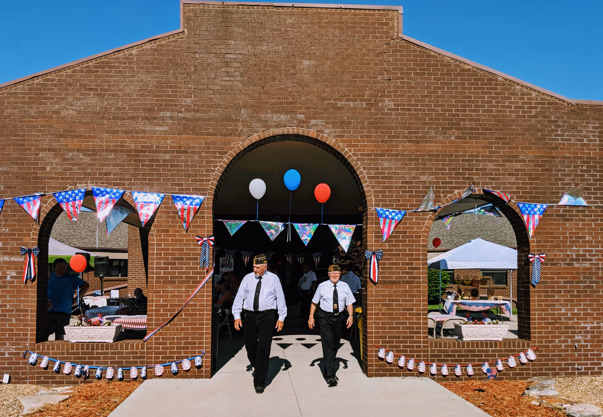Two men in white shirts, black pants, and black hats walk out of a brick building decorated with American flag-themed bunting and balloons in red, white, and blue. There are tents and tables with red and white checkered tablecloths visible inside and outside the building under a clear blue sky.