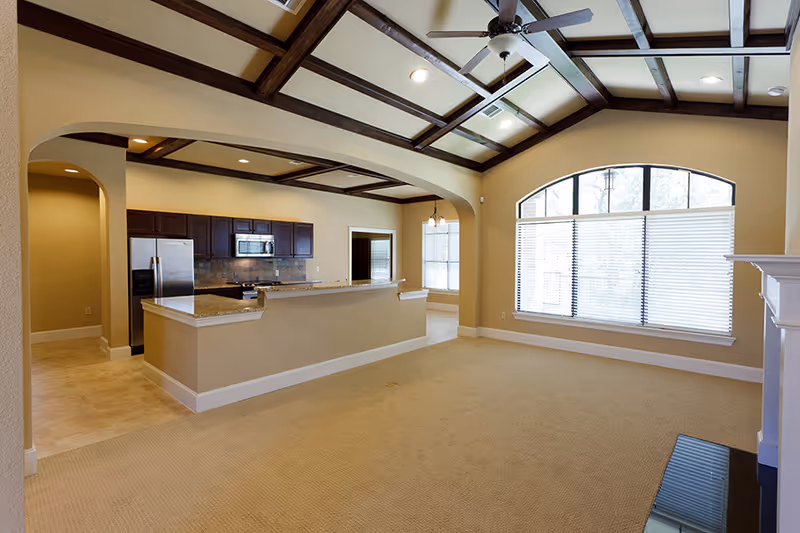 Spacious open-concept living area with beige carpet, a large arched window with blinds, and a kitchen featuring dark wood cabinets, stainless steel appliances, and a granite countertop bar. The ceiling has exposed wooden beams and recessed lighting.