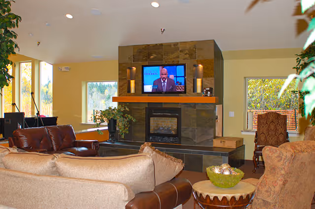 Cozy lounge with sofas and armchairs facing a stone fireplace topped by a wall-mounted TV, with windows letting in natural light.