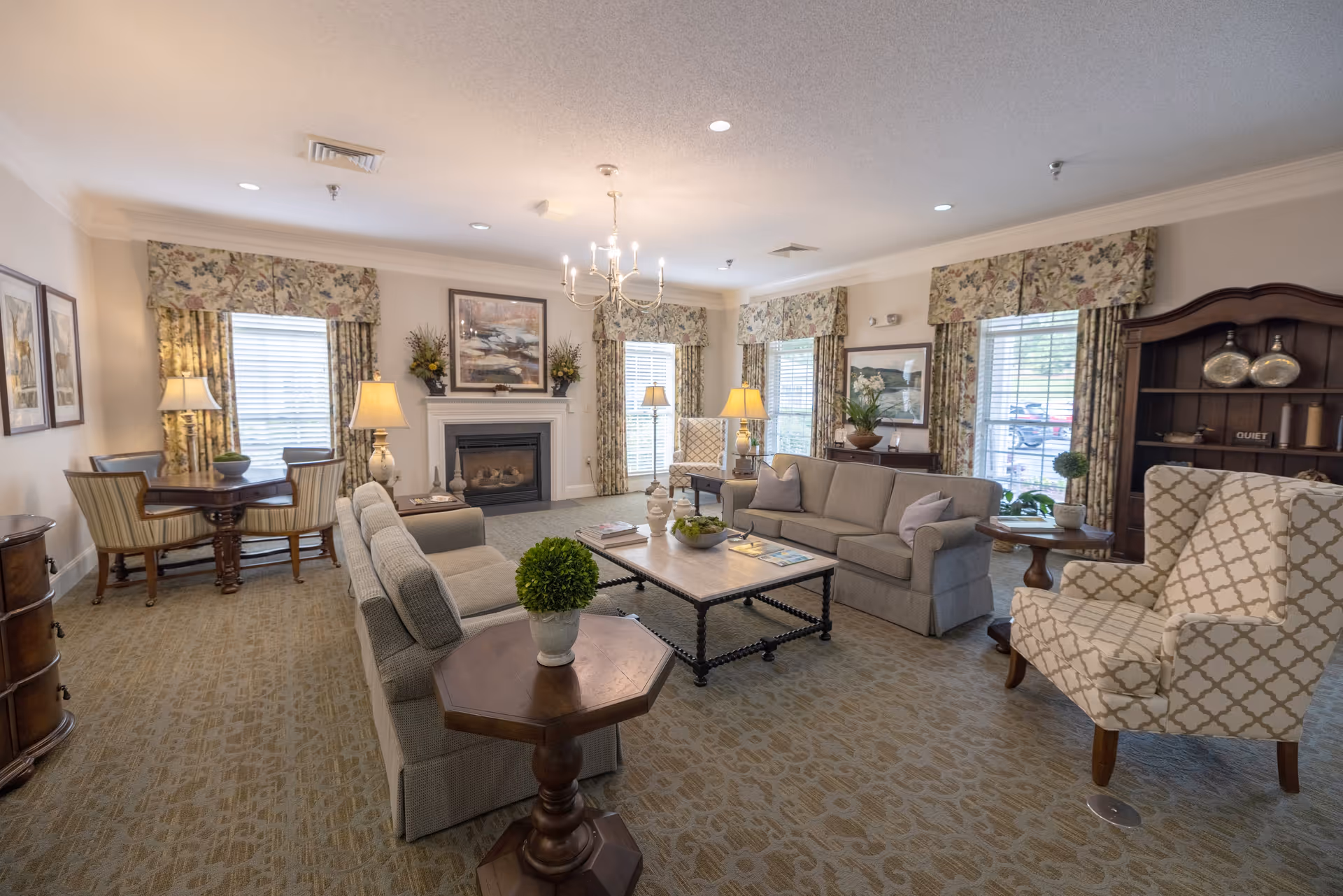 A cozy living room with two gray sofas, a patterned armchair, a wooden coffee table with decorative items, and a fireplace with floral arrangements on the mantle. The room has floral curtains on the windows, a chandelier hanging from the ceiling, and a small dining table with four chairs in the corner.