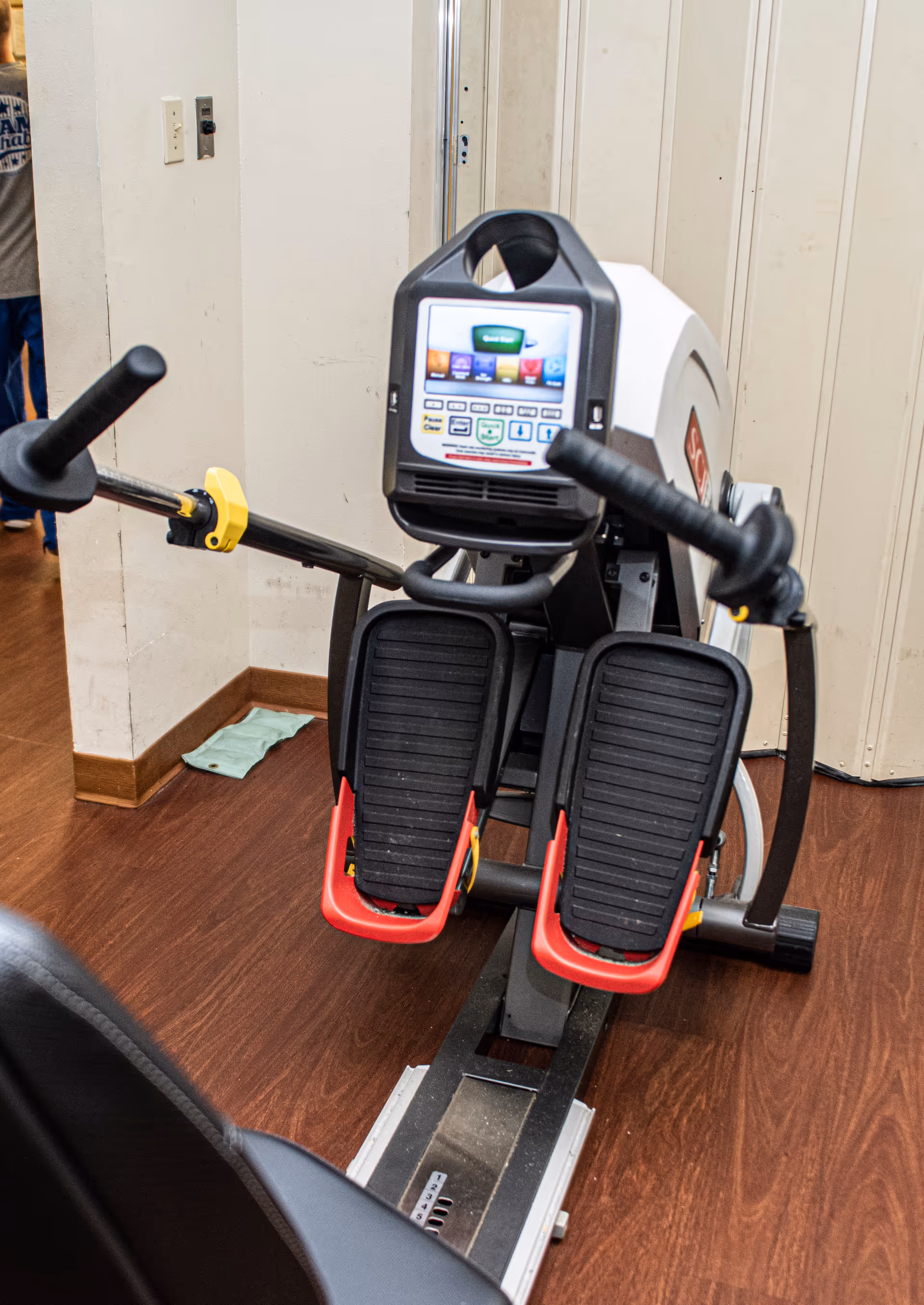 A close-up view of a rowing exercise machine with foot pedals and a digital display screen in a room with wooden flooring and beige walls.