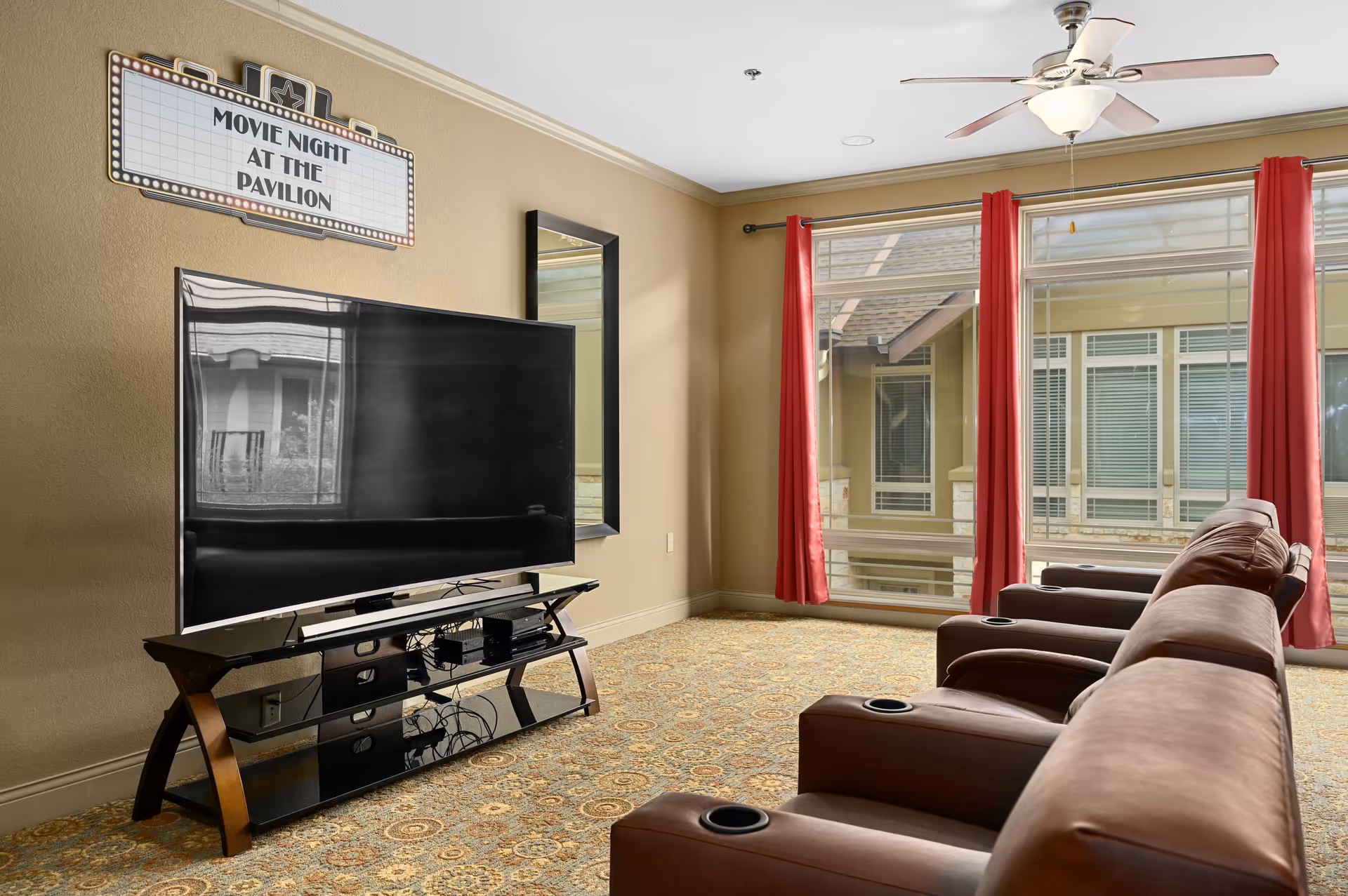 A cozy lounge area with a large flat-screen TV on a stand, three brown recliner chairs with cup holders facing the TV, large windows with red curtains letting in natural light, a ceiling fan, and a sign on the wall that reads 'Movie Night at The Pavilion'.