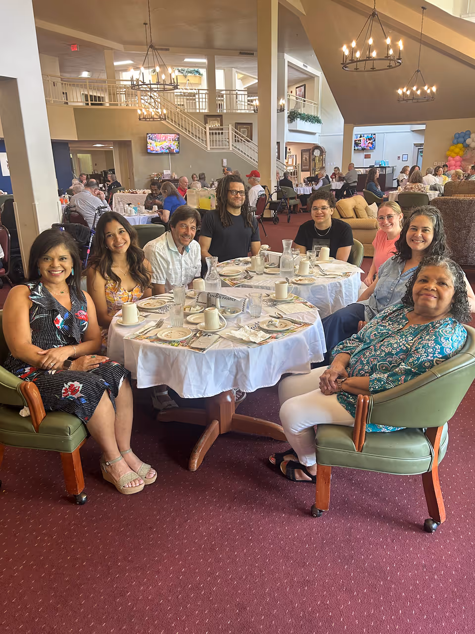 A group of eight people sitting around a round dining table set with plates, cups, and utensils in a spacious, well-lit dining area with high ceilings and chandeliers. Other people are seated at tables in the background, and there is a staircase and television visible in the room.