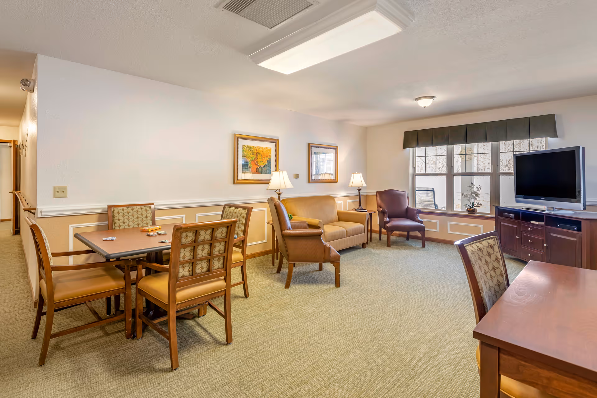A well-lit living room area in a senior living facility featuring a small dining table with four chairs, a beige loveseat, two armchairs, two table lamps, framed artwork on the wall, a large window with a valance, and a flat-screen TV on a wooden cabinet.