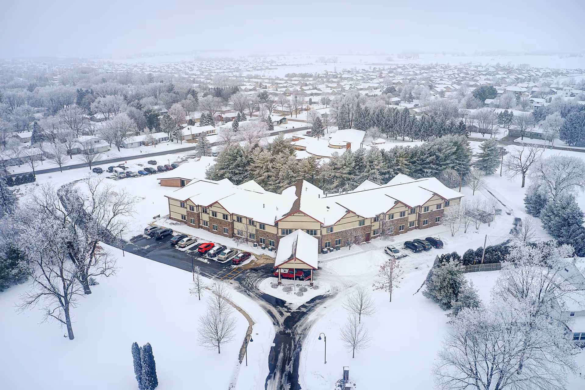 Aerial view of The Heights at Evansville Manor covered in snow, showing a large two-story building with a parking lot partially cleared of snow, surrounded by snow-covered trees and residential houses in the background.