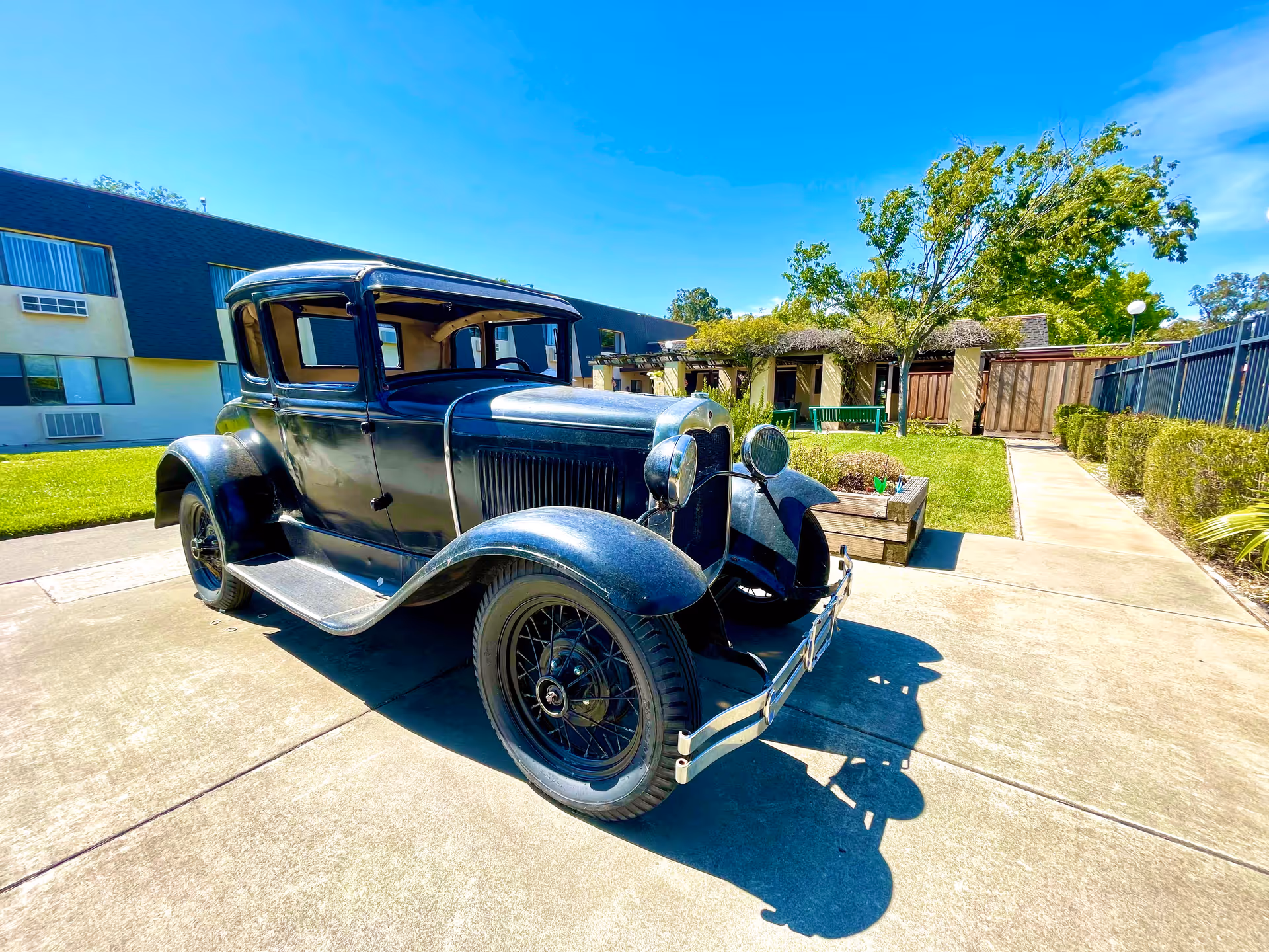 A vintage black car parked on a concrete driveway in front of a residential building with green grass, trees, and a clear blue sky.