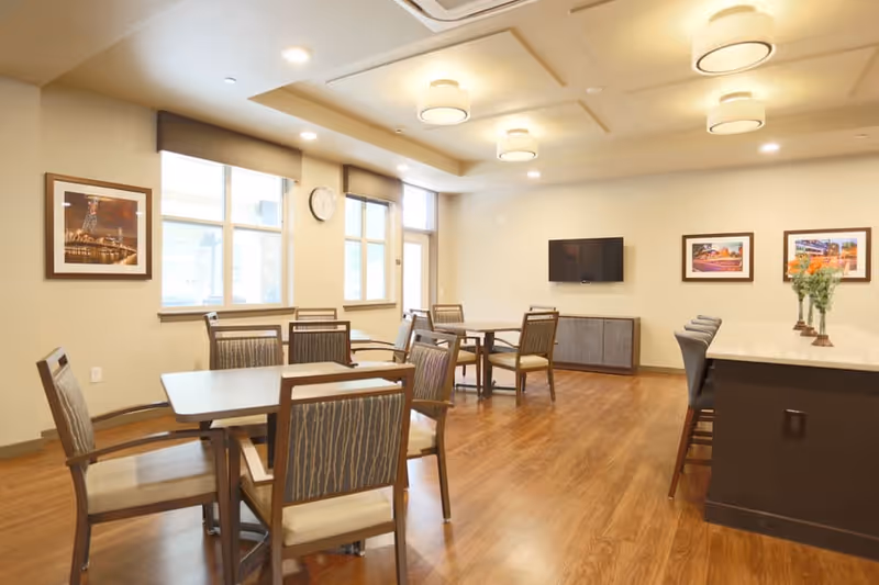 A bright and spacious dining area in a senior living facility featuring several wooden tables with cushioned chairs, large windows letting in natural light, a wall-mounted flat screen TV, framed artwork on the walls, and a countertop with bar stools and flower vases.