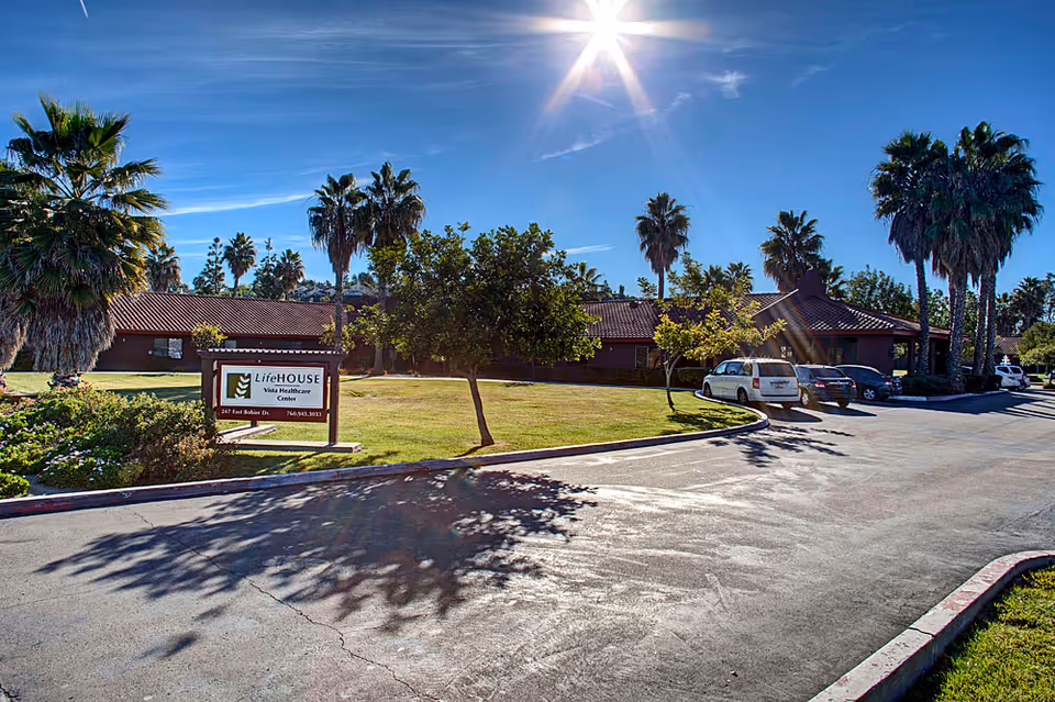 Exterior view of a single-story healthcare facility building with a tiled roof, surrounded by palm trees and a well-maintained lawn under a bright sunny sky. There is a sign in front that reads 'LifeHOUSE Vista Healthcare Center' along with an address and phone number. Several cars are parked along the driveway.