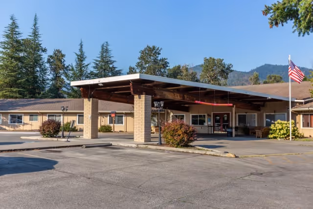 Exterior view of Highland House Nursing & Rehabilitation Center showing a single-story building with a large covered entrance supported by brick columns, surrounded by trees and mountains in the background, with an American flag on a flagpole near the entrance.