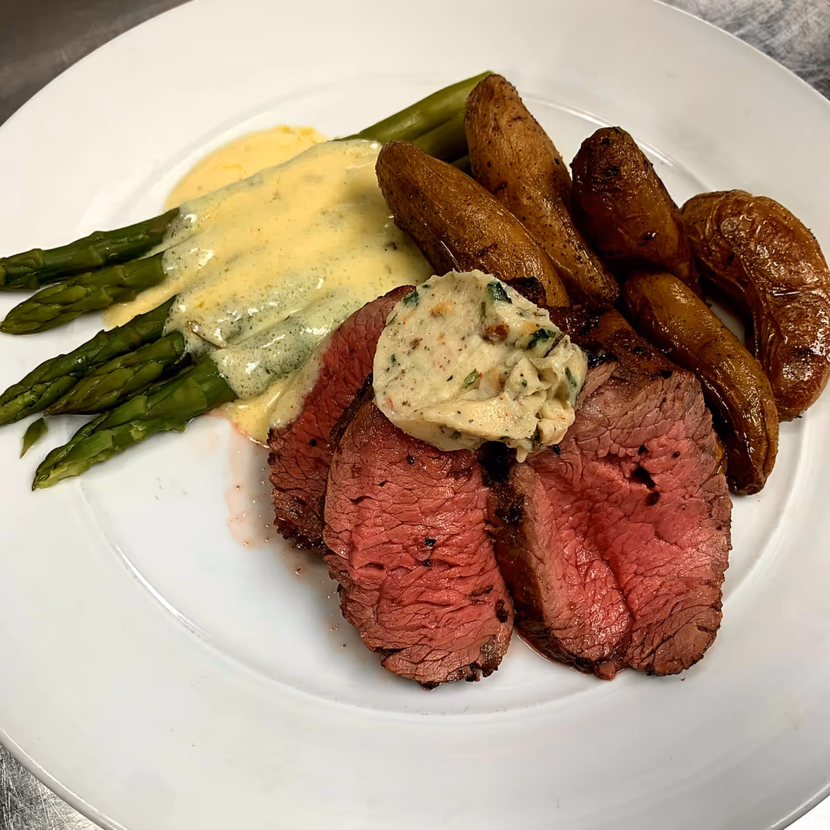 A plate of food featuring three slices of medium-rare roast beef topped with a dollop of herb butter, roasted fingerling potatoes, and steamed asparagus with a creamy sauce.