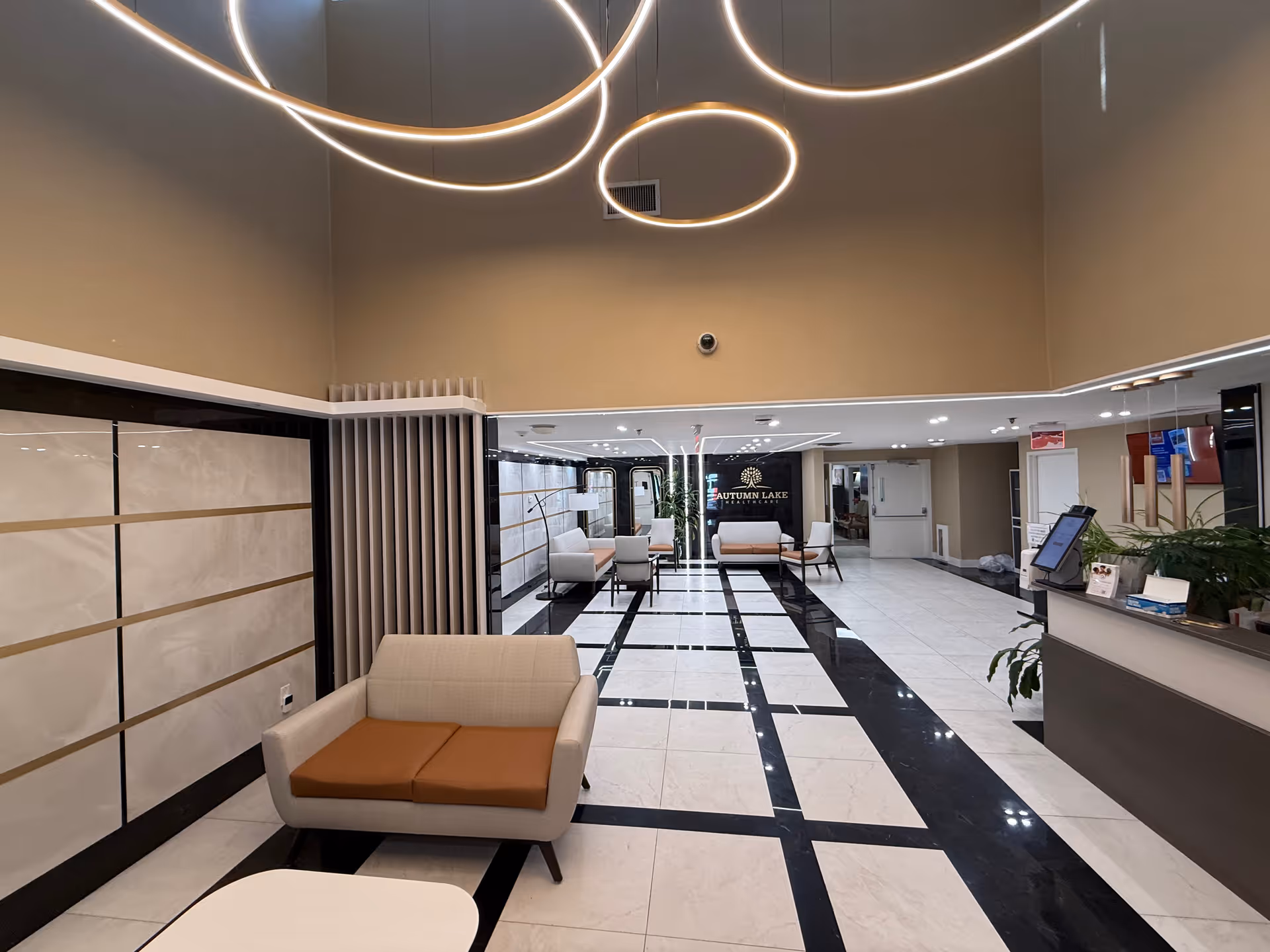 A modern and spacious lobby area with high ceilings and circular light fixtures hanging from above. The floor has a black and white checkered tile pattern. There are several beige and brown cushioned chairs arranged for seating. A reception desk is visible on the right side with some plants and a computer monitor. The back wall features a sign that reads 'Autumn Lake Healthcare'.