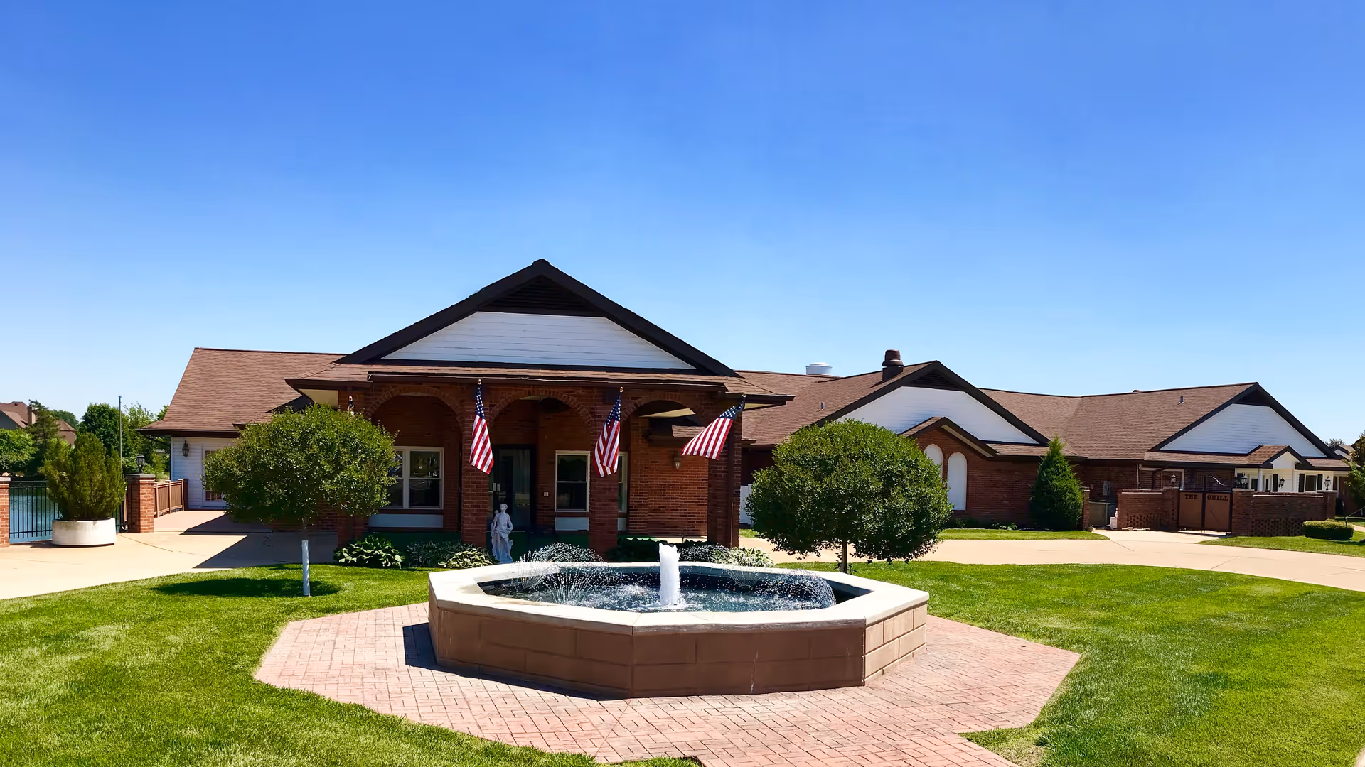 Front of Mari De Villa Nursing Home showing a brick building with American flags, a central fountain, and a manicured lawn under a clear blue sky.