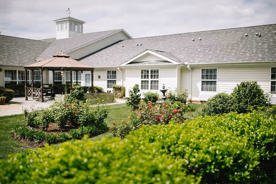 Outdoor garden area at Spring Arbor of Raleigh featuring well-maintained green bushes, flowering plants, a gazebo with seating, and a white building with multiple windows in the background under a cloudy sky.