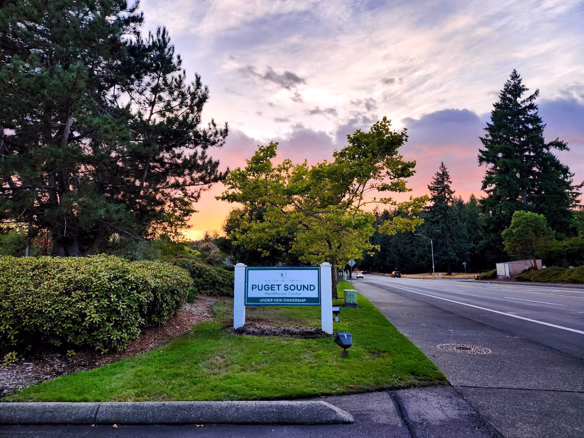 Entrance sign for Puget Sound Care by a tree-lined road at sunset.
