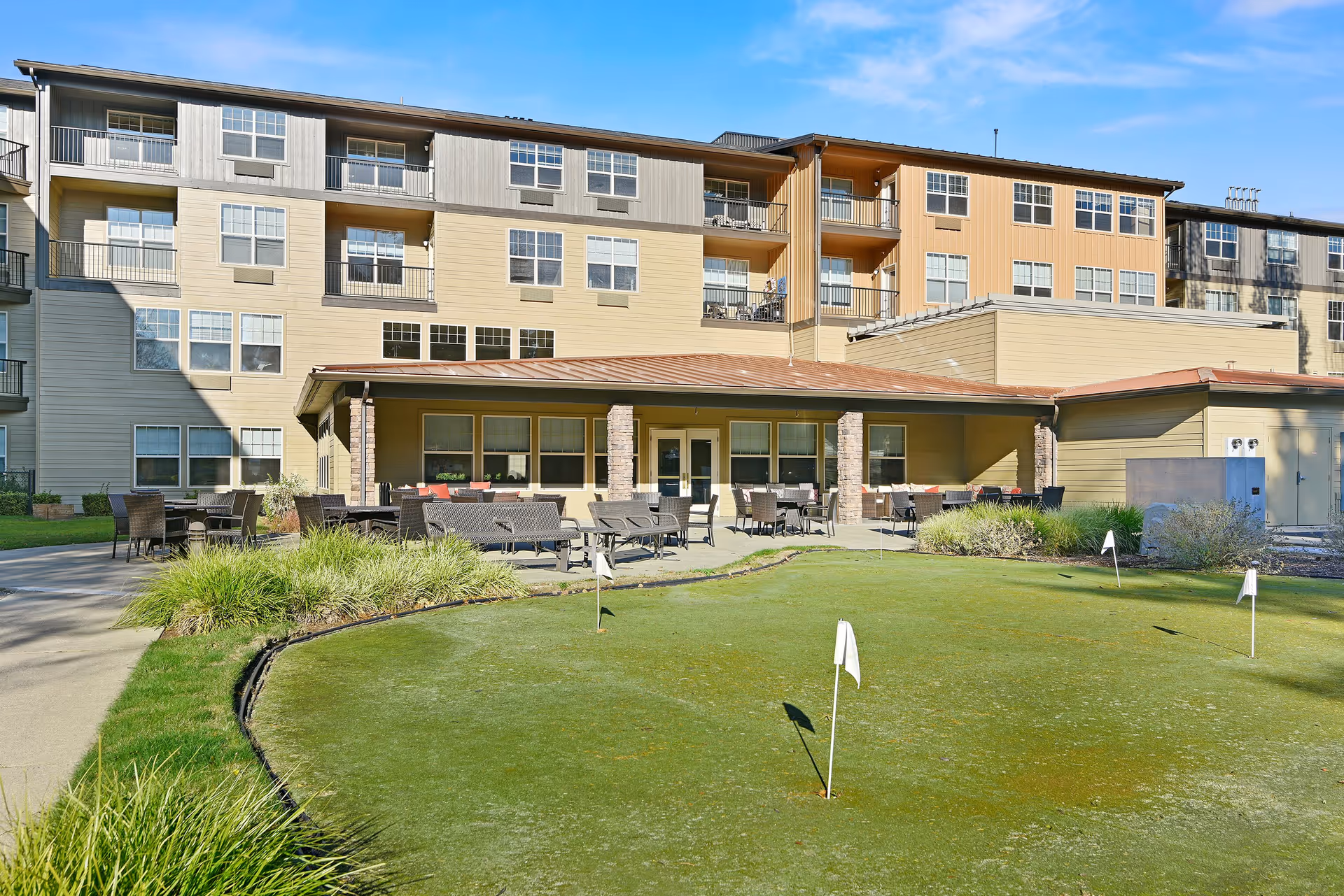 Outdoor view of a senior living facility with a putting green in the foreground and a patio area with tables and chairs in front of a multi-story building under a clear blue sky.
