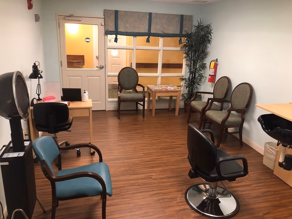 A small salon-style room with barber chairs, a hooded hair dryer, waiting chairs, and a desk on wood floors.