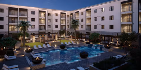 Evening view of a senior living facility courtyard with a large swimming pool surrounded by lounge chairs, fire pits, palm trees, and string lights. The multi-story building with balconies and windows encloses the courtyard.