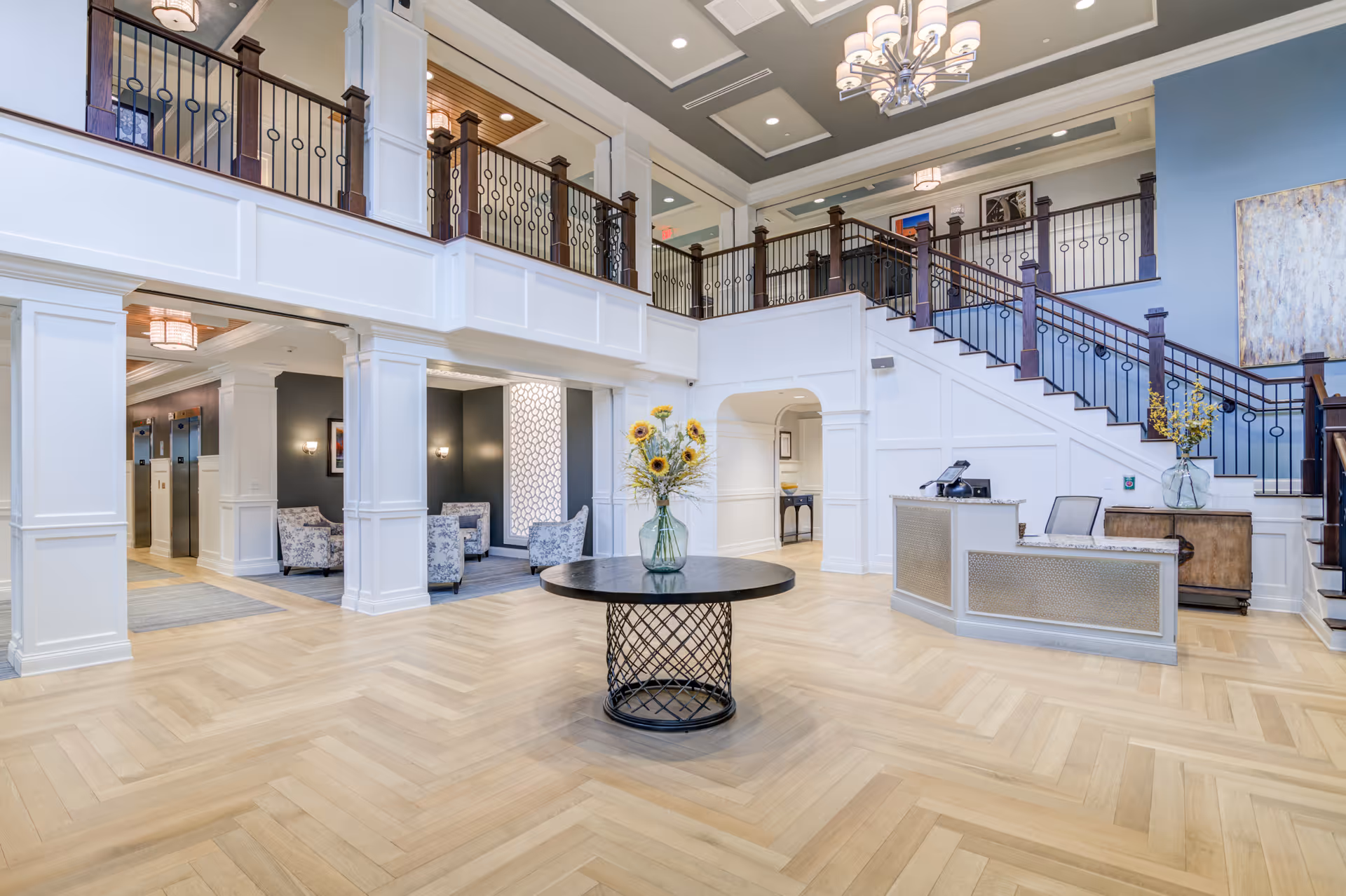Spacious and elegant lobby area of an assisted living facility with a round table featuring a vase of sunflowers in the center, a reception desk to the right, and a staircase leading to an upper level with decorative railings. The space has high ceilings with chandeliers, light wood flooring, and seating areas visible in the background.