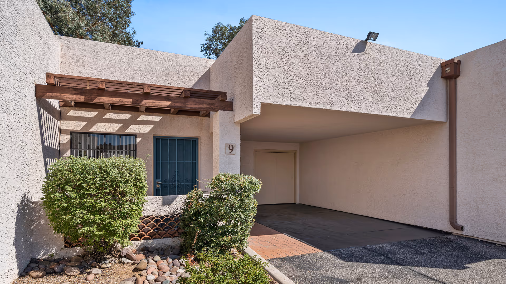 Front exterior of a beige stucco unit with a covered carport, wood pergola, entry door marked '9' and trimmed shrubs.