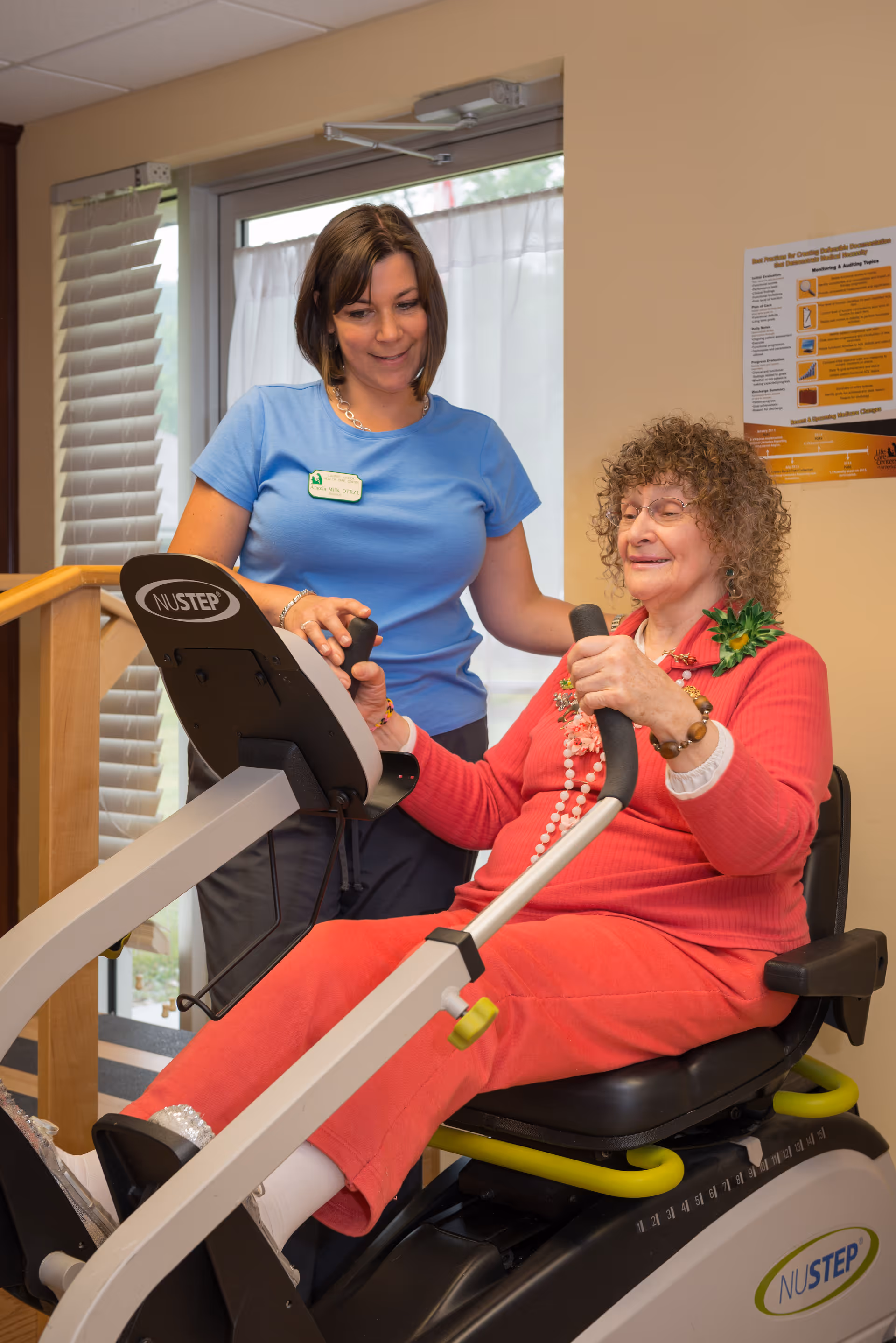 An elderly woman with curly hair wearing a red outfit is seated on a NuStep exercise machine, holding the handles and smiling. A female caregiver or fitness instructor in a blue shirt stands beside her, offering support and encouragement. The setting appears to be an indoor exercise or rehabilitation area with a window and informational poster on the wall.