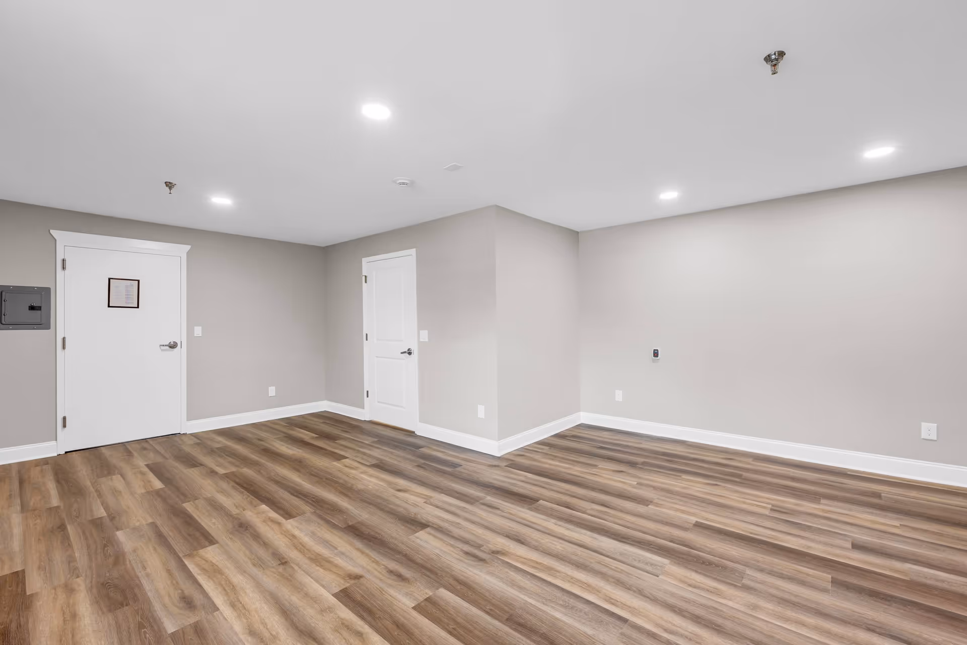 Empty room with light gray walls, two white doors, recessed ceiling lights, and wood-patterned vinyl flooring.