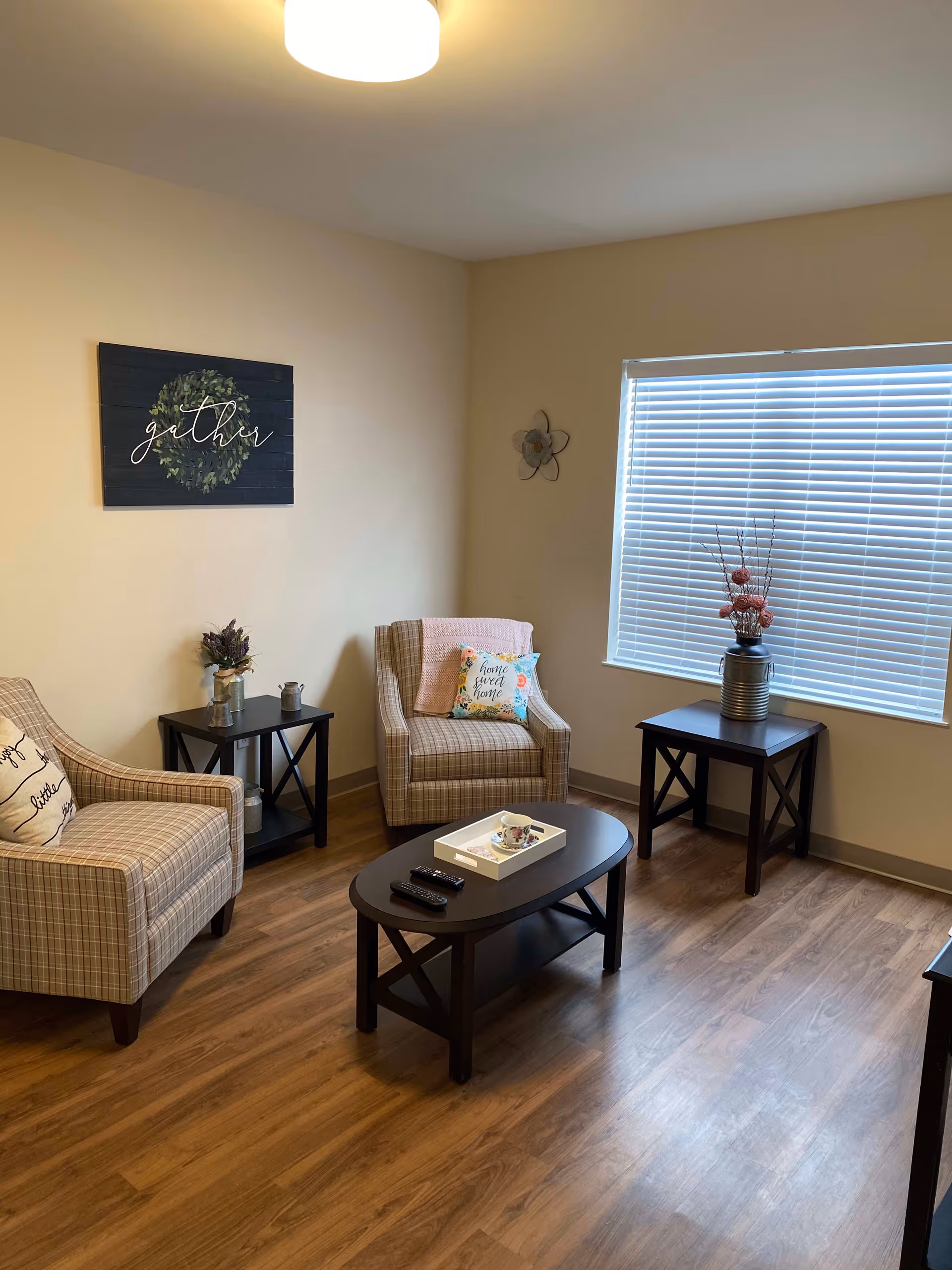 A cozy living room with two plaid armchairs, each with decorative pillows and a pink throw blanket on one. There are two dark wooden side tables, one with a vase of flowers and the other with a decorative arrangement. A matching dark wooden coffee table holds a white tray with a teacup and two remote controls. The room has wood flooring, beige walls, a large window with white blinds, and a ceiling light. A wall decoration with the word 'gather' is hung on the wall.